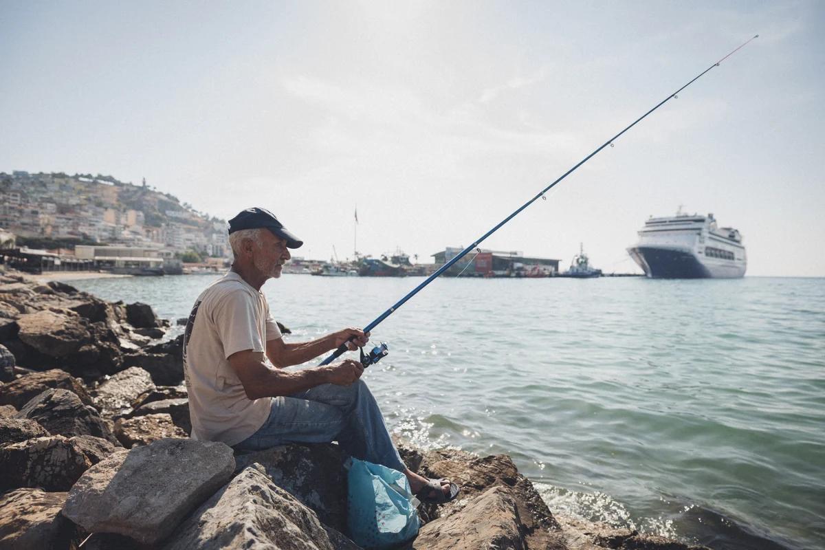 A fisherman seated on coastal rocks with a ship in the distance